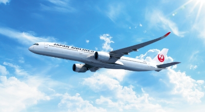 A Japan Airlines airplane flying through a bright blue sky with scattered clouds.