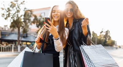 Two young women smiling and holding shopping bags while looking at a smartphone outdoors on a sunny day.