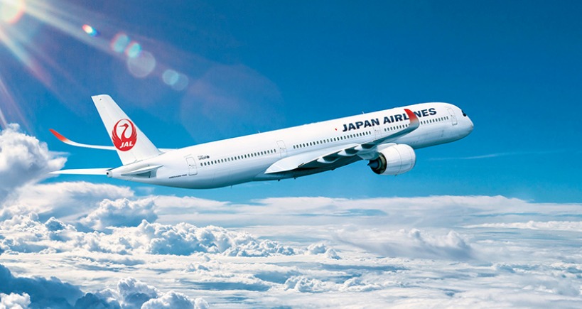 A Japan Airlines airplane flying through a bright blue sky with scattered clouds.